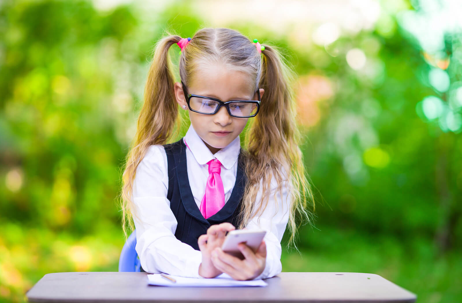 A young girl is sitting at the desk in myopia control glasses and typing on her phone. 