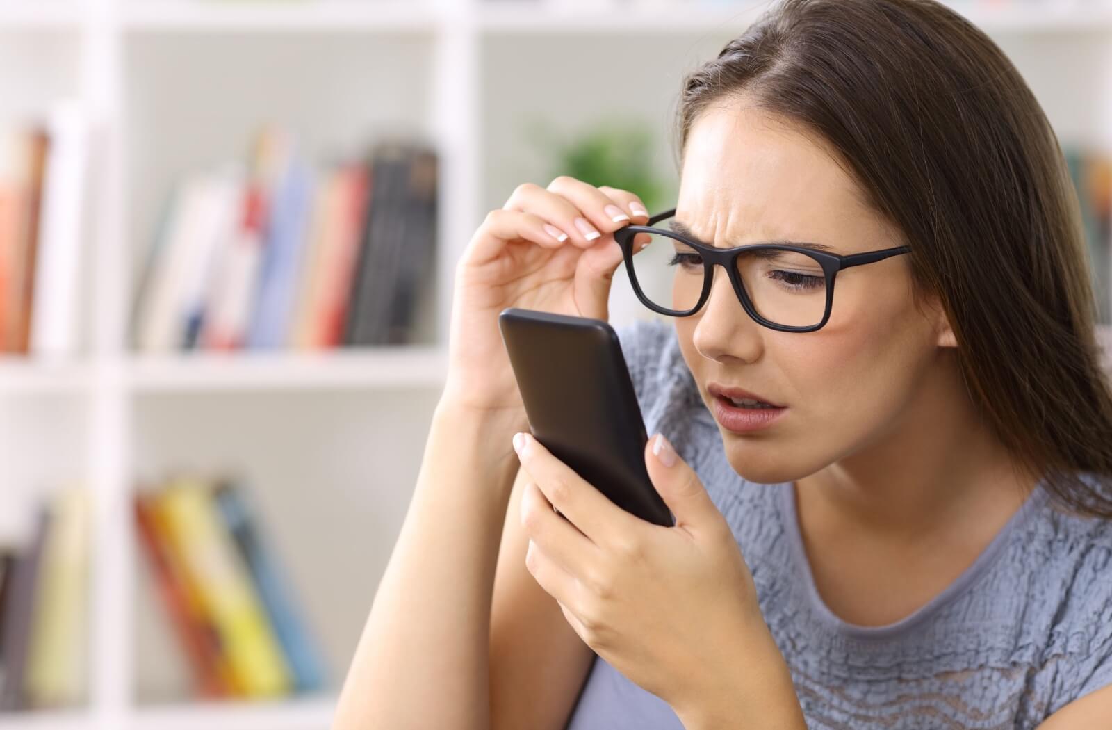 A woman holding her phone close to her face while adjusting her glasses to see its contents better.