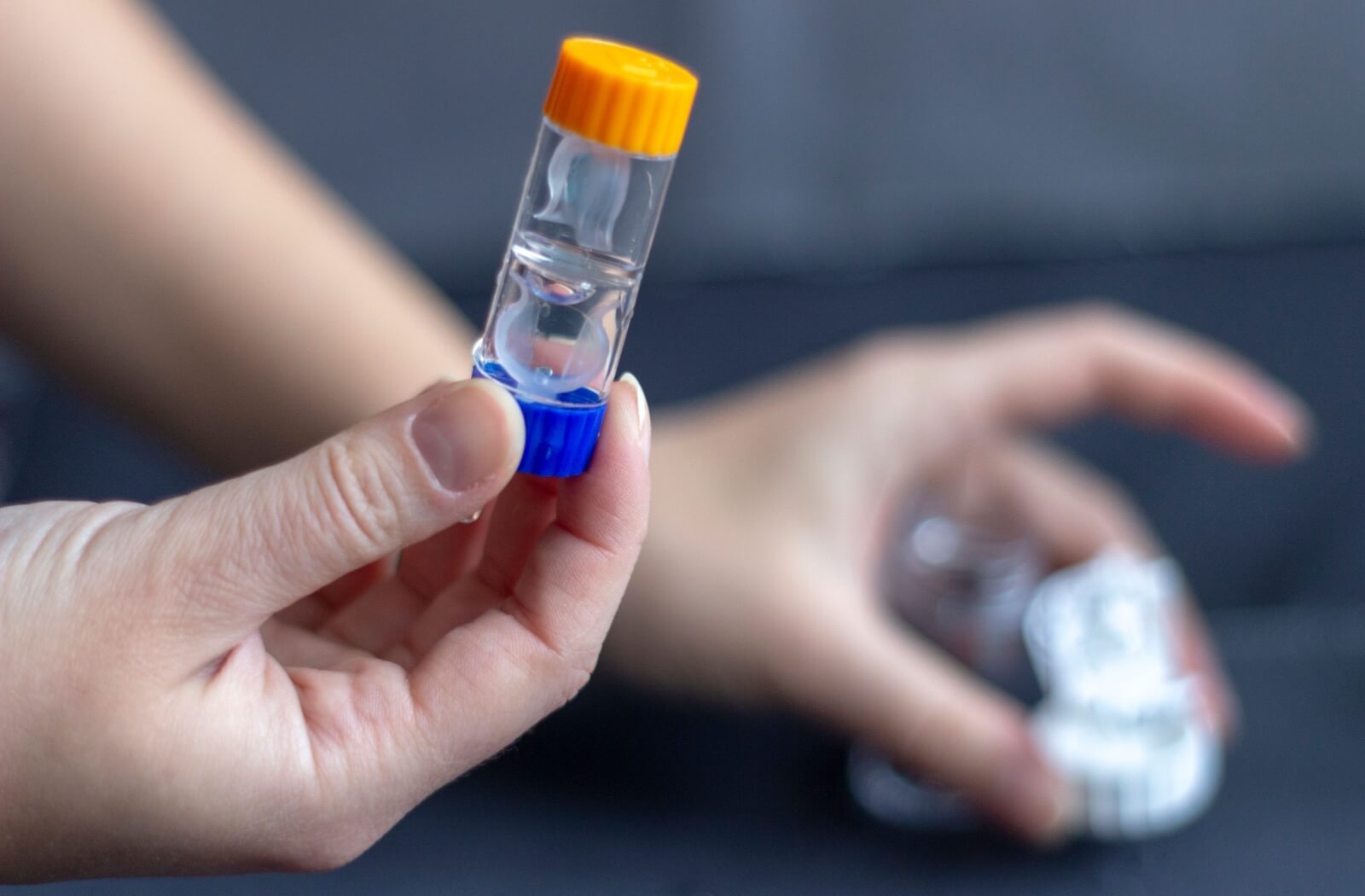 A close-up of a pair of contact lenses in a clear vial, immersed in cleaning solution. A person holds the vial at one end, preparing to open it.