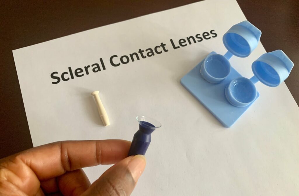 A contact lens case rests against a piece of paper that reads "Scleral Contact Lenses", while a hand in the foreground holds a scleral lens suction tool.