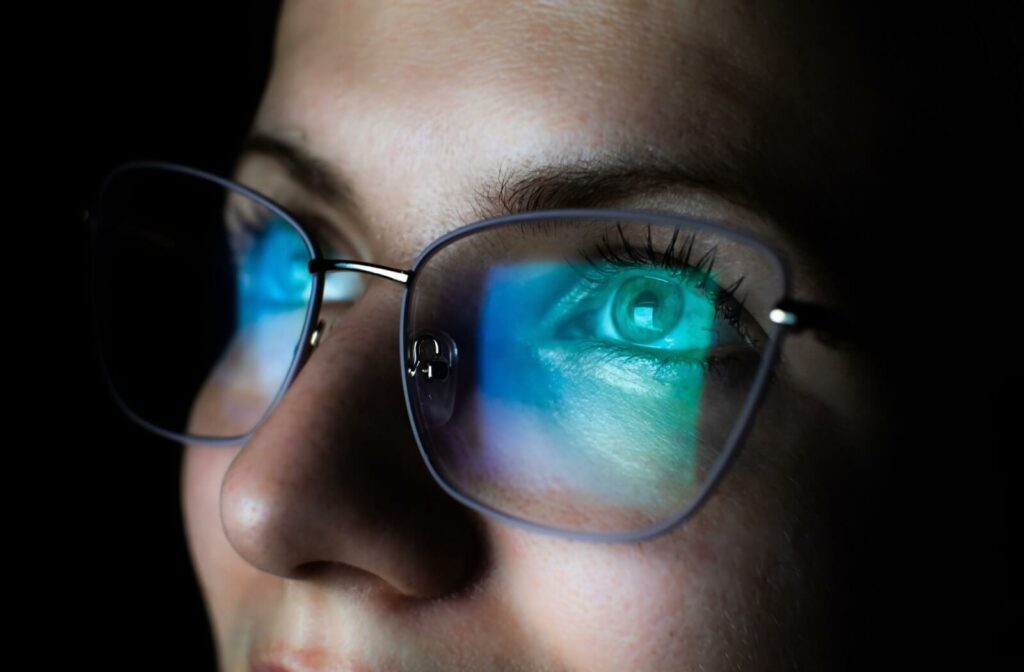 A close-up of a person wearing blue light glasses that are reflecting a computer screen in a dark room.