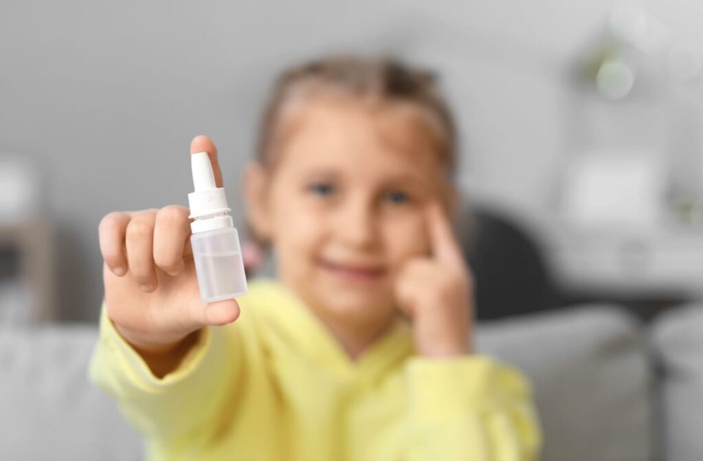 A close-up in-focus image of a young child in a yellow sweater smiling and holding up a bottle of eye drops.