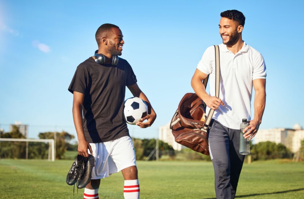 Two adults walking on a soccer pitch; one carries cleats and a soccer ball and the other carries a water bottle and bag.
